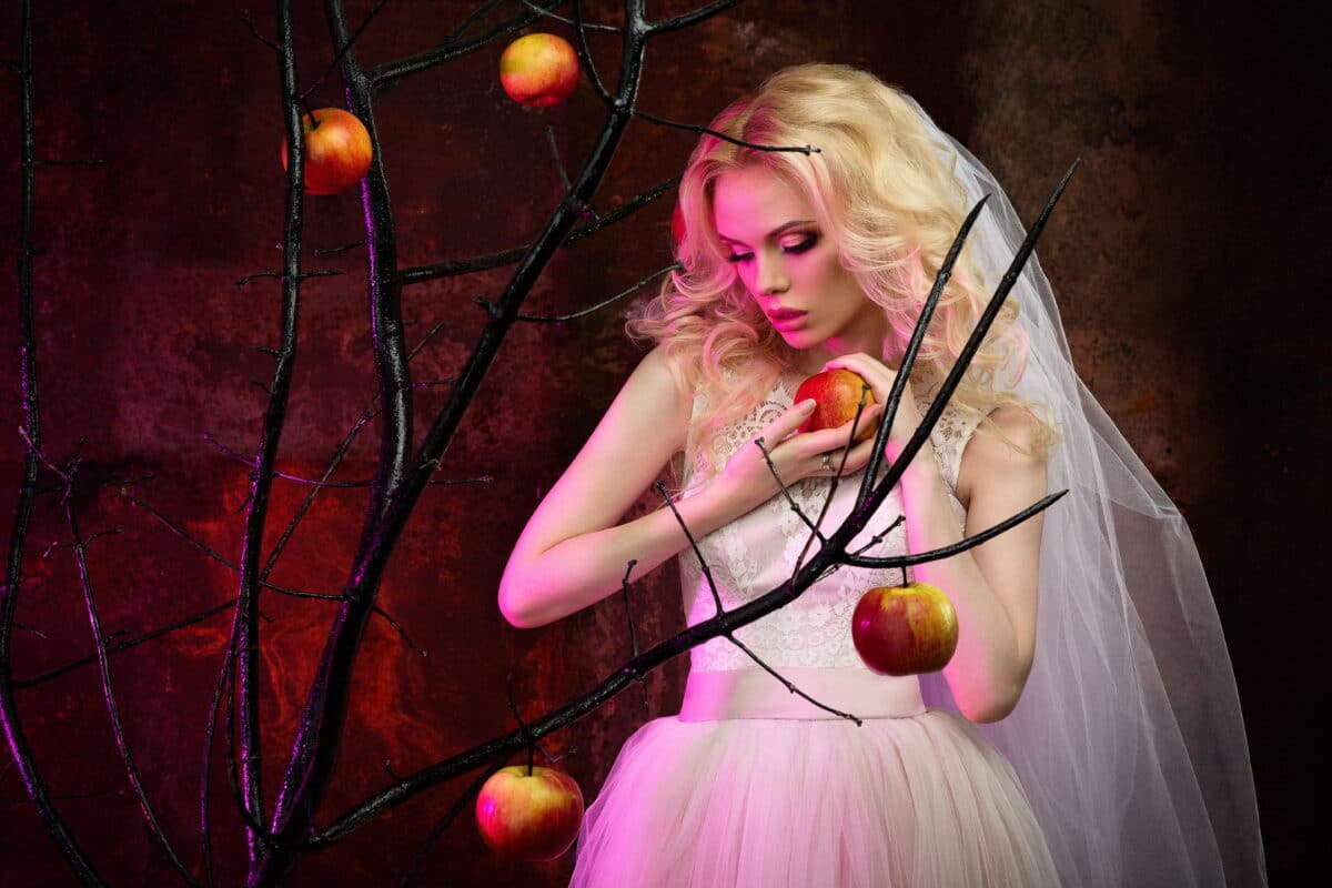 beautiful blonde girl in a white dress standing by a leafless tree with red apples
