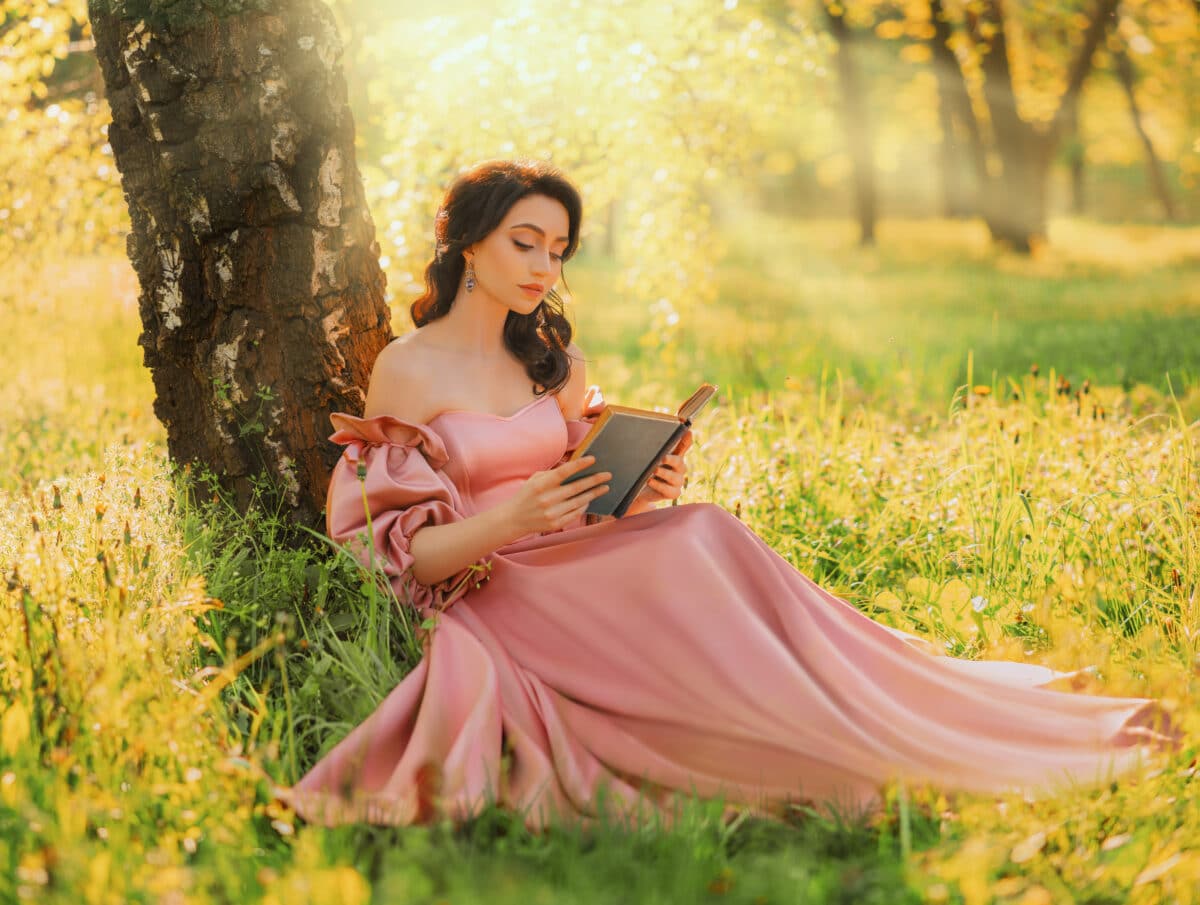 Fantasy woman sits under tree holding romantic book in hands reading novel. Pink long vintage dress. Fairy princess girl in garden summer nature green grass magic sun rays light.