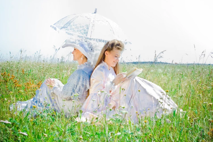 Two beautiful women in vintage dress sitting back to back reading outdoor on the grass.