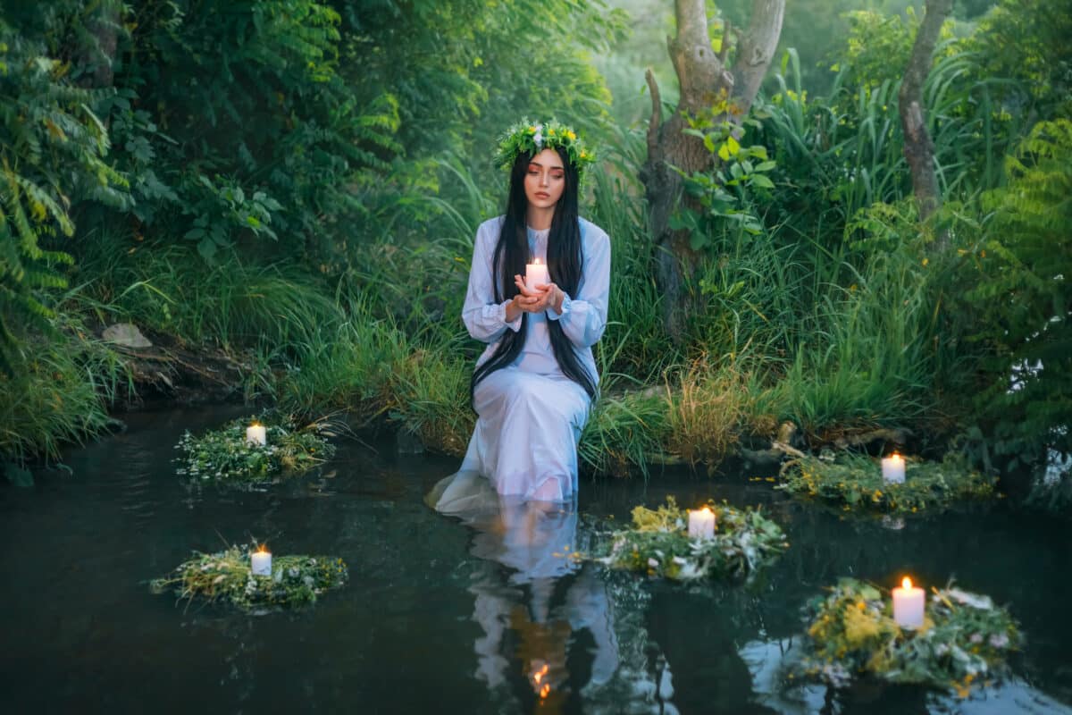fantasy pagan witch slavic woman holding candle in hand. Divination summer tradition. Girl Mermaid nymph sits on banks river. White vintage nightgown dress. summer nature green grass trees. Long hair
