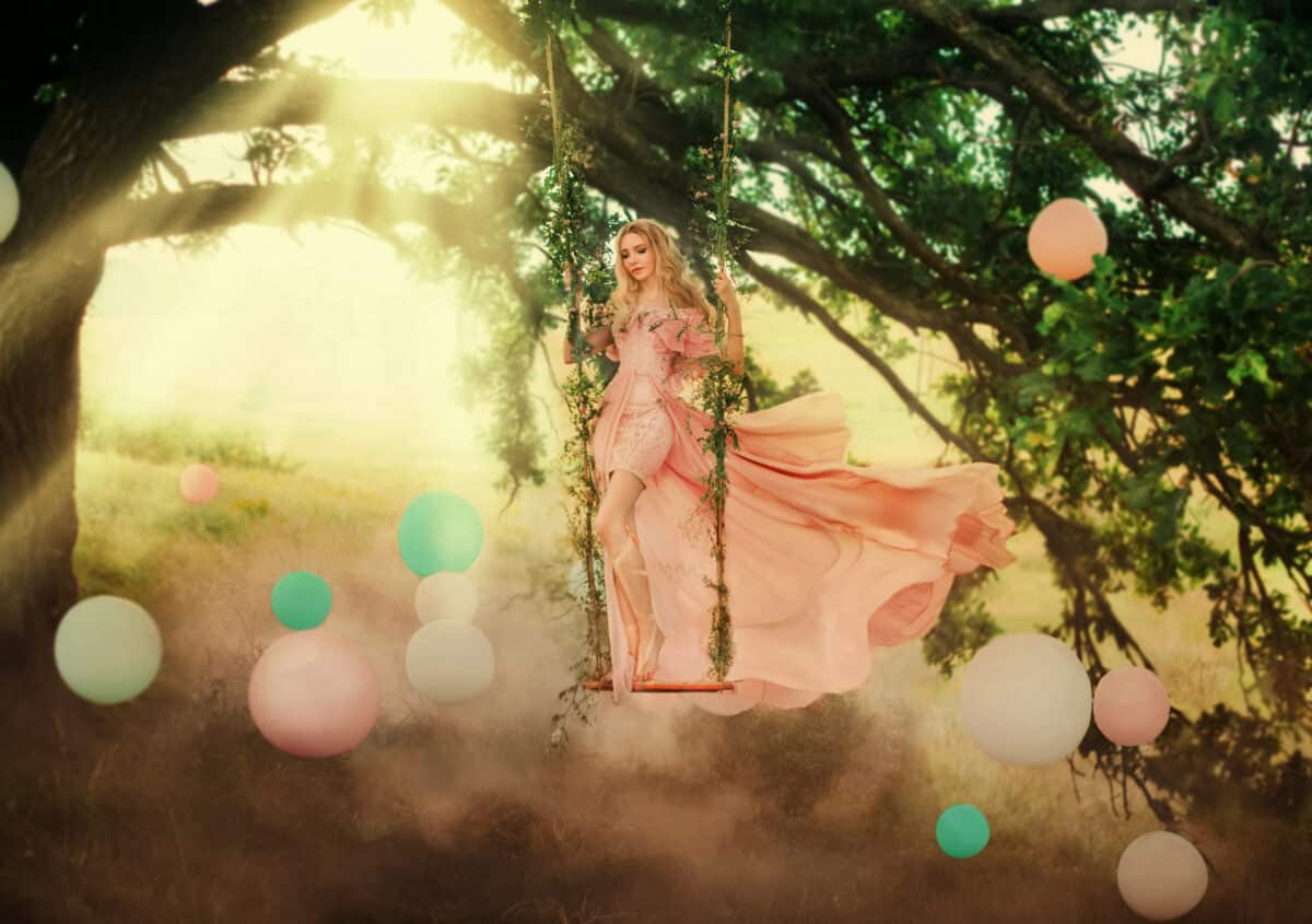 Art photo happy playful fantasy blonde woman princess having fun swinging on swing. Wood background, colored smoke balloons sun light. Pink silk dress flies in wind. Positive emotions smile on face