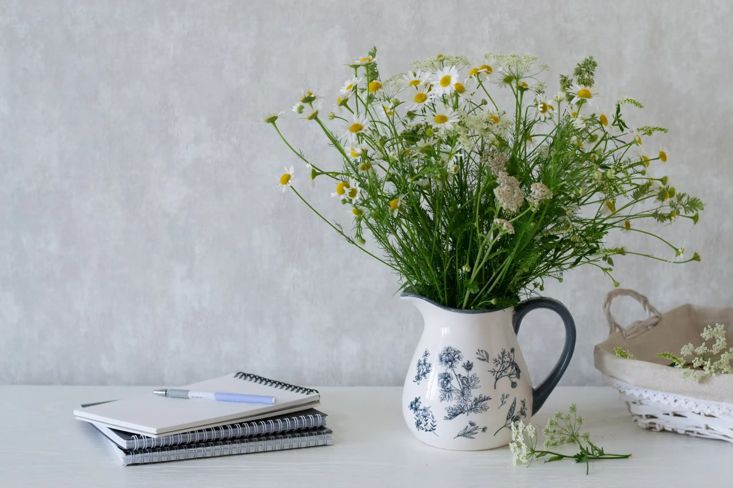 Vase of white daisies next to stacked notebooks on white desk.
