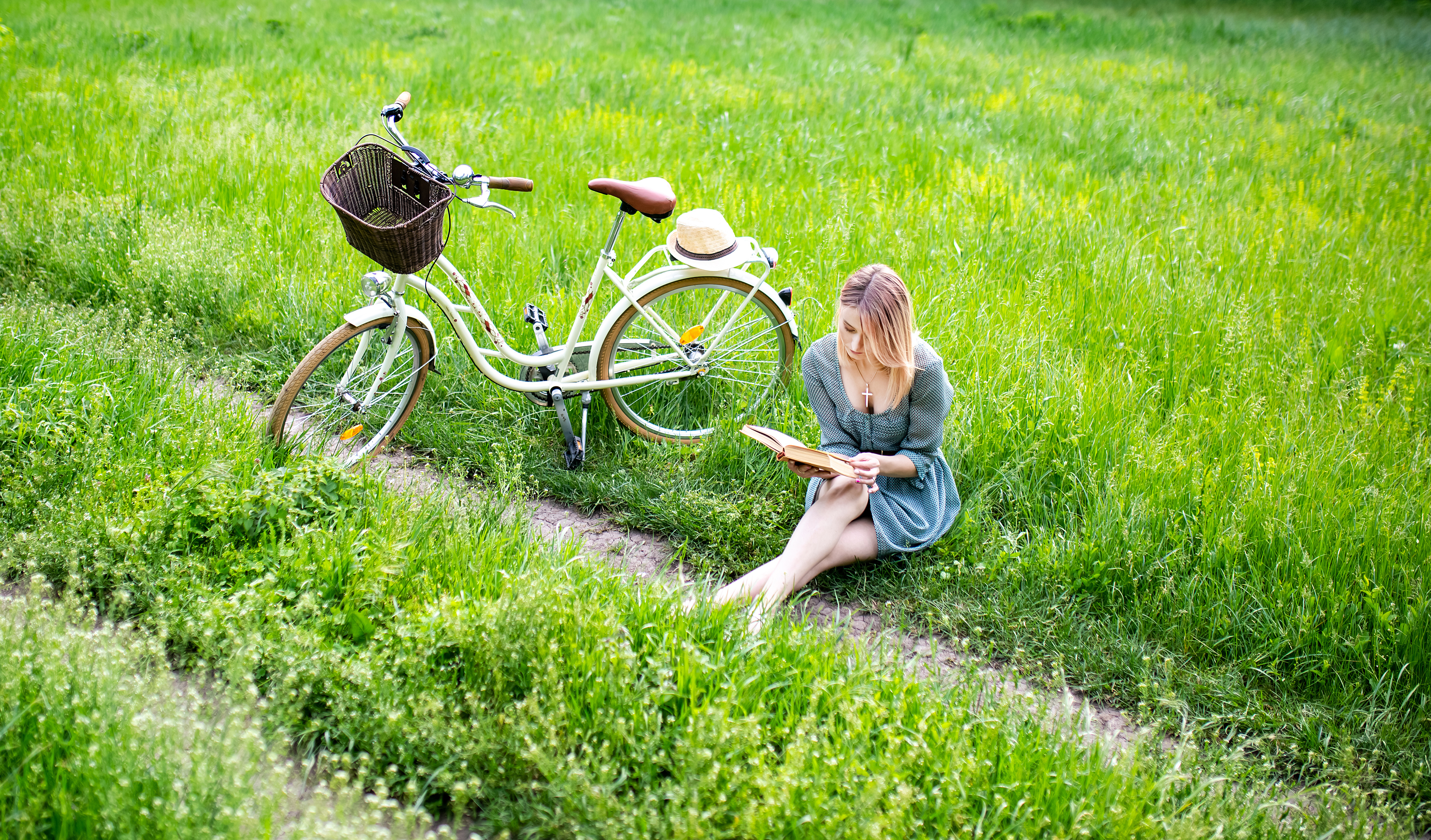  A girl is reading a book next to her bicycle out in the green field.