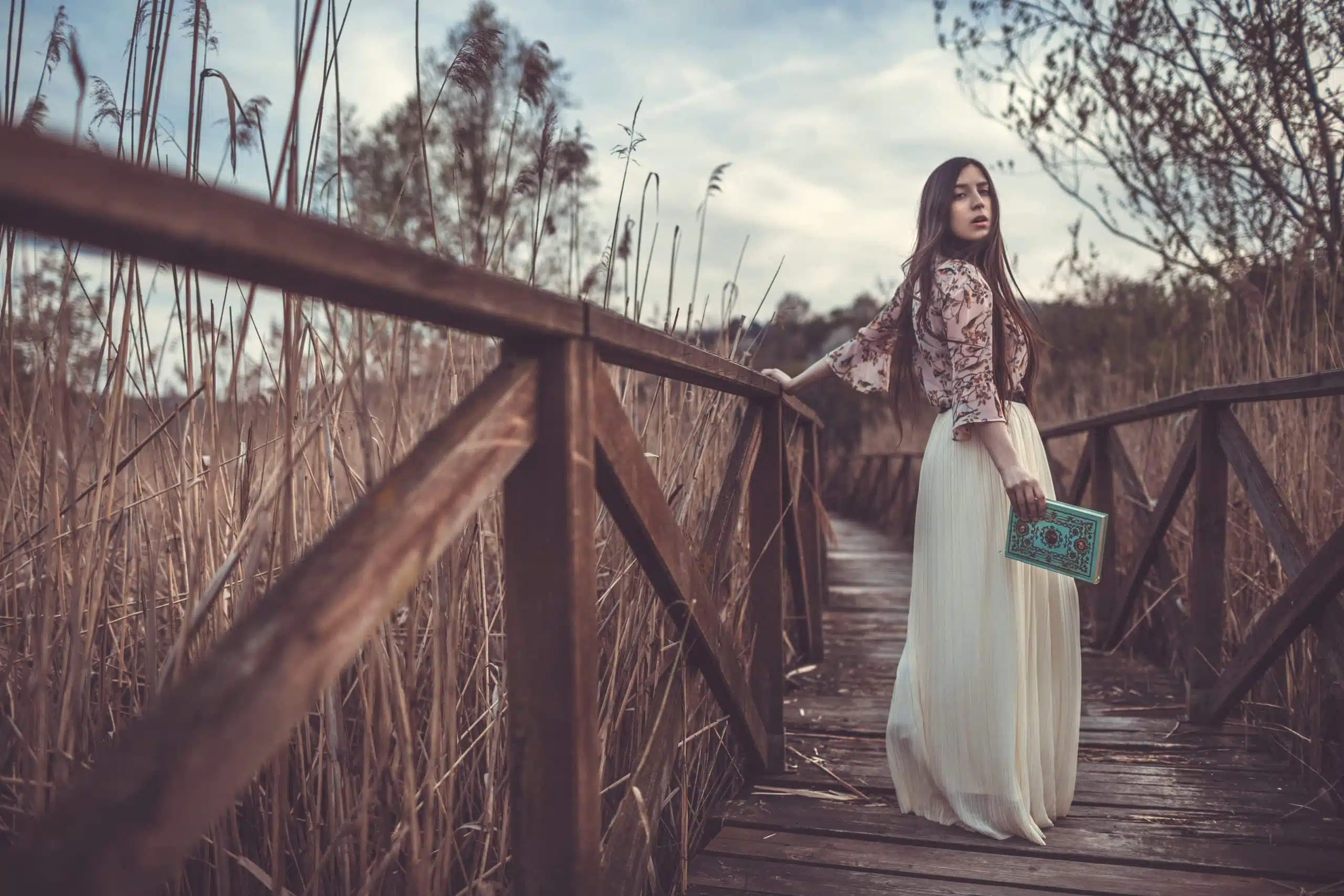 Beautiful young woman holding a book, walking on a wooden bridge.