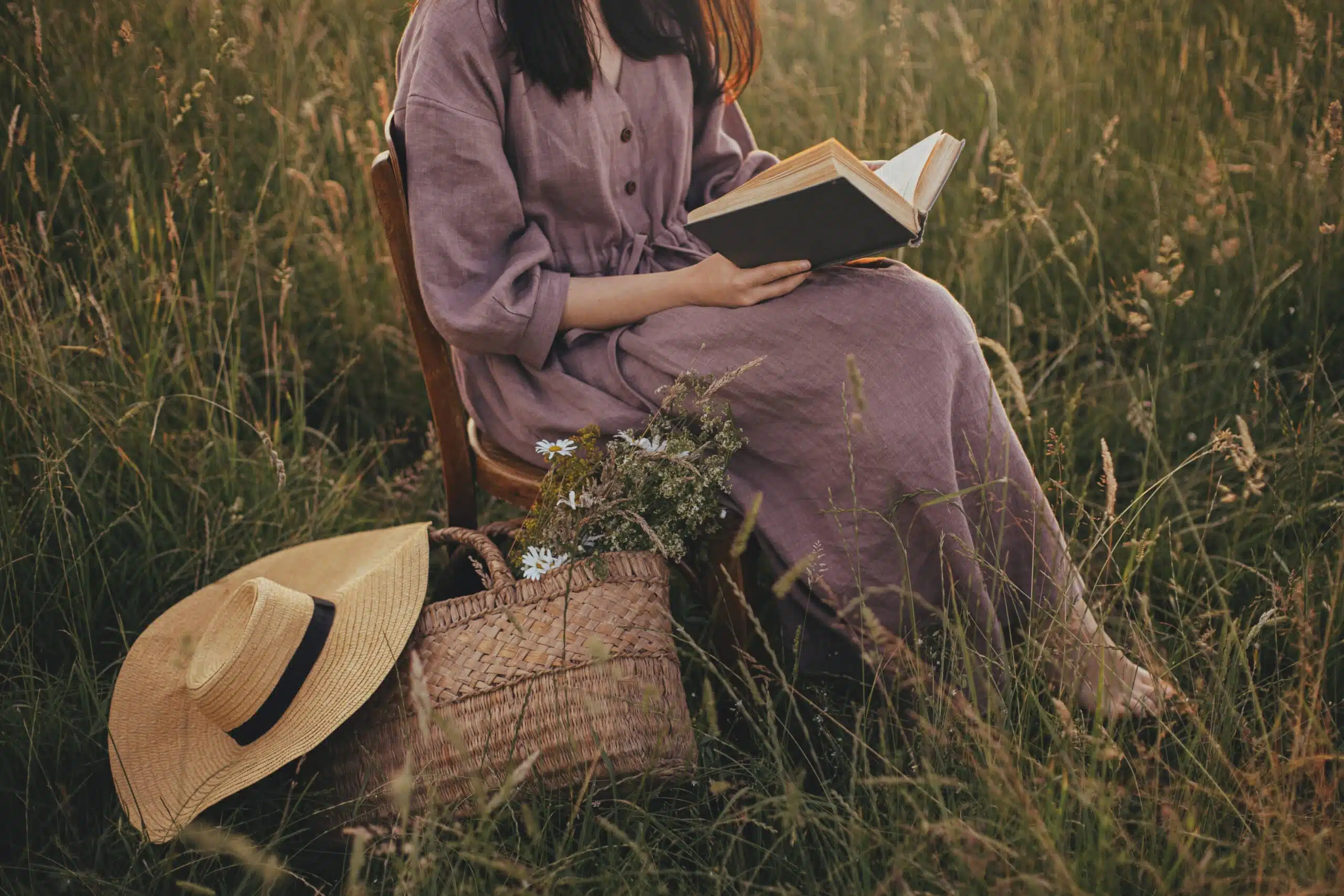 Beautiful woman in linen dress with book and basket of flowers sitting in the field.