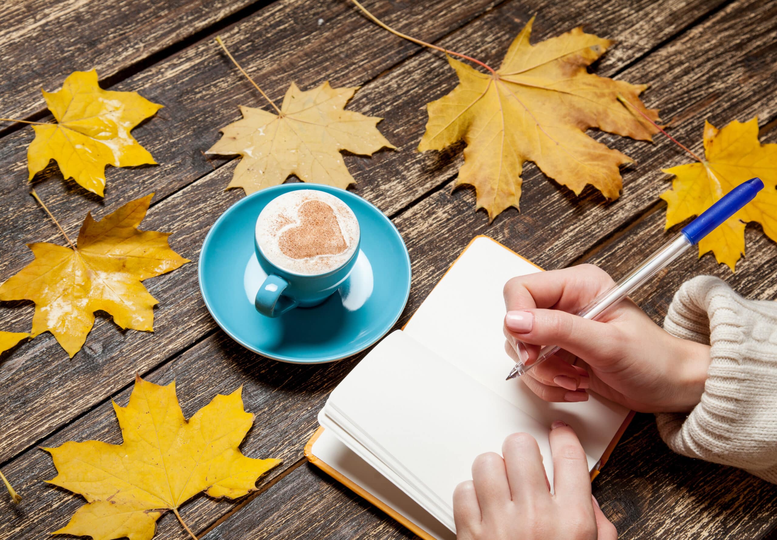 Female hand writing in notebook near cup of coffee on wood table with maple leaves