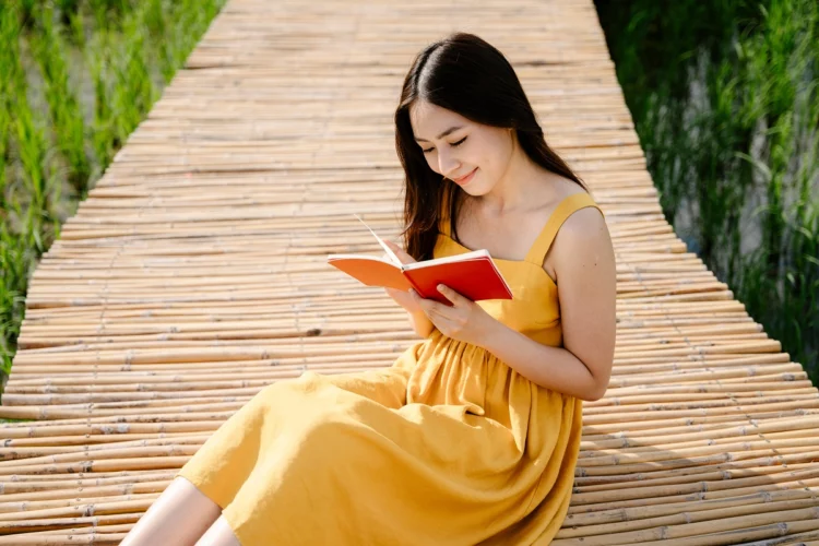 Asian woman in yellow dress reading red book on wooden foot bridge.