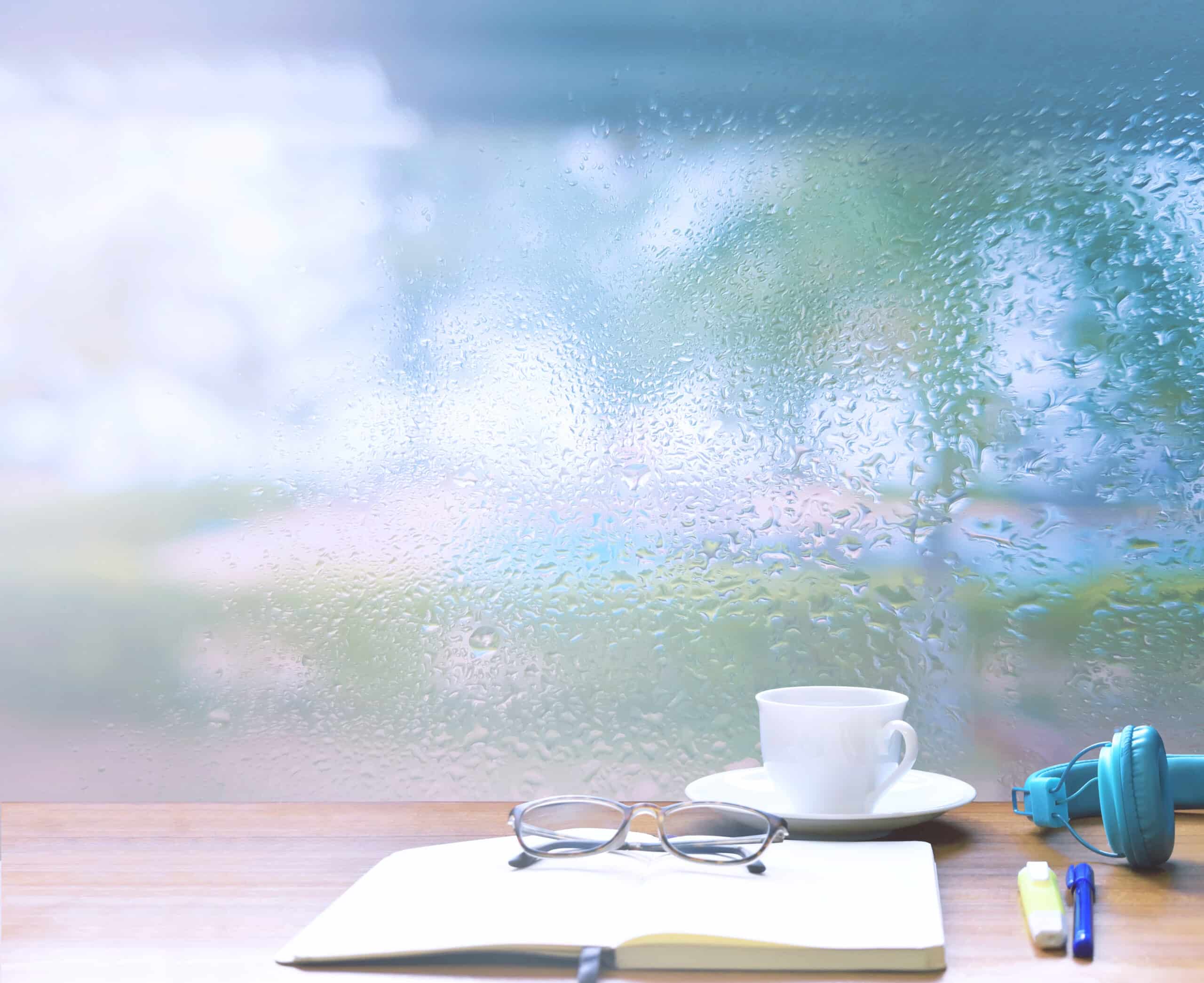 water drops forming on the glass window, open blank notebook, glasses, and a cup on windowsill