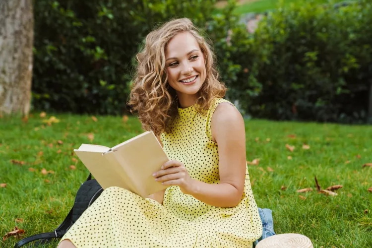 pleased blonde woman smiling and reading book while sitting on the grass.