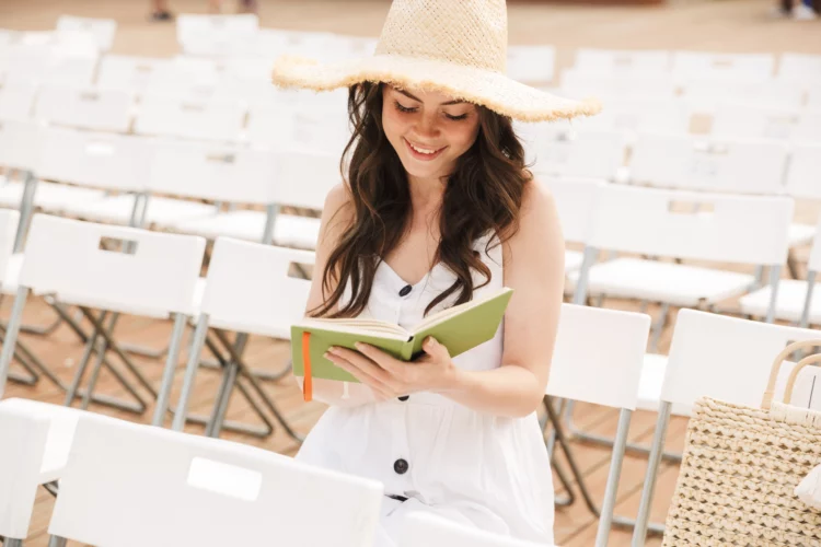 alluring woman wearing straw hat smiling and holding diary book while sitting in a chair at a park