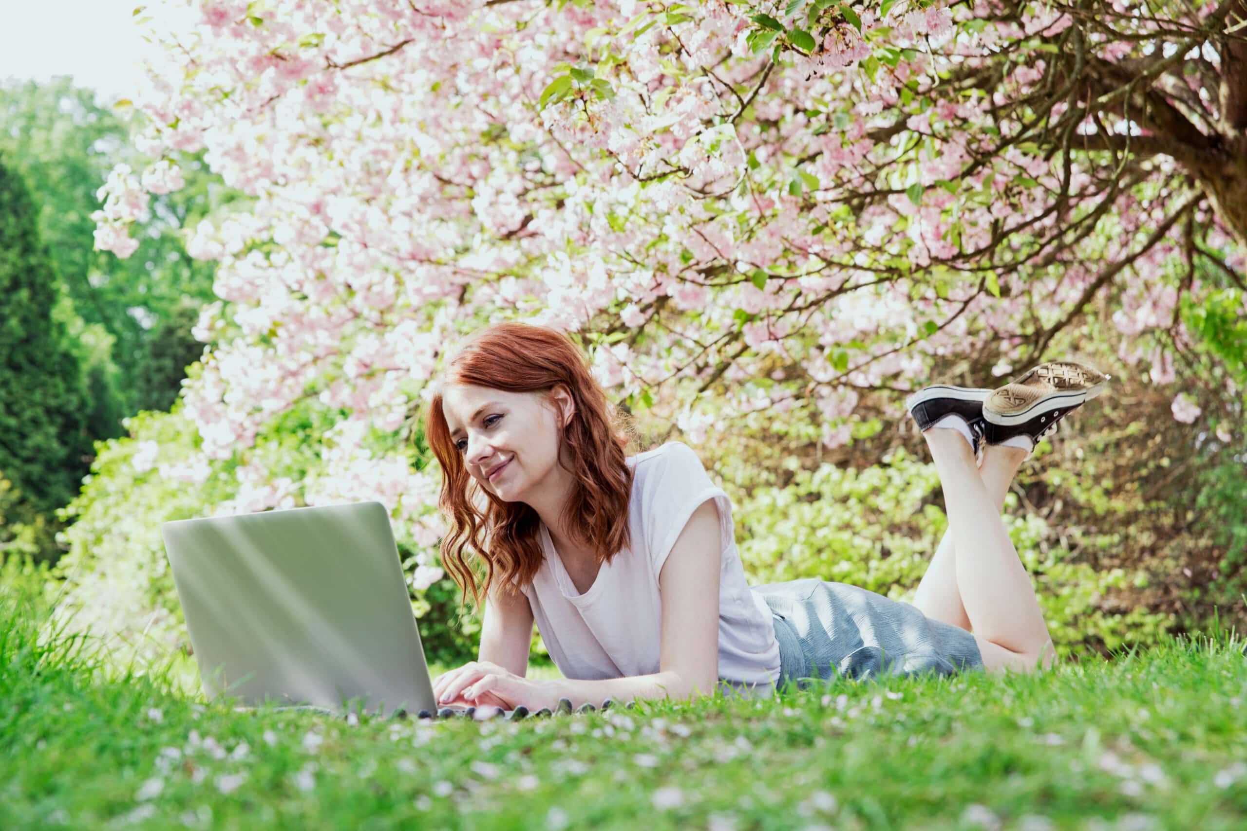 smiling redhead girl working on her laptop in cherry blossom tree garden
