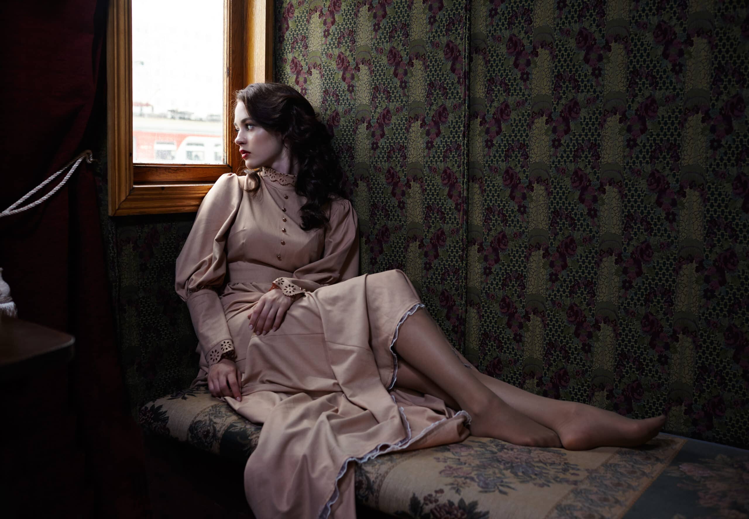Young alluring woman in beige vintage dress of early 20th century sitting on a couch while looking out the window.