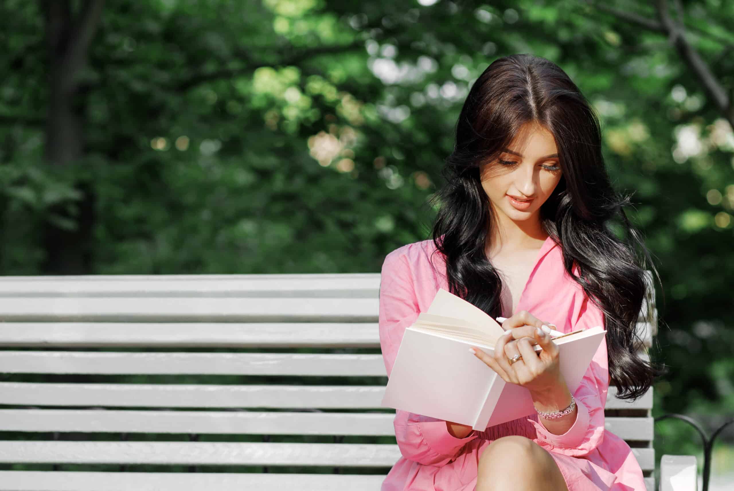 Beautiful young woman in pink dress, writing in notebook sitting on bench outdoor.