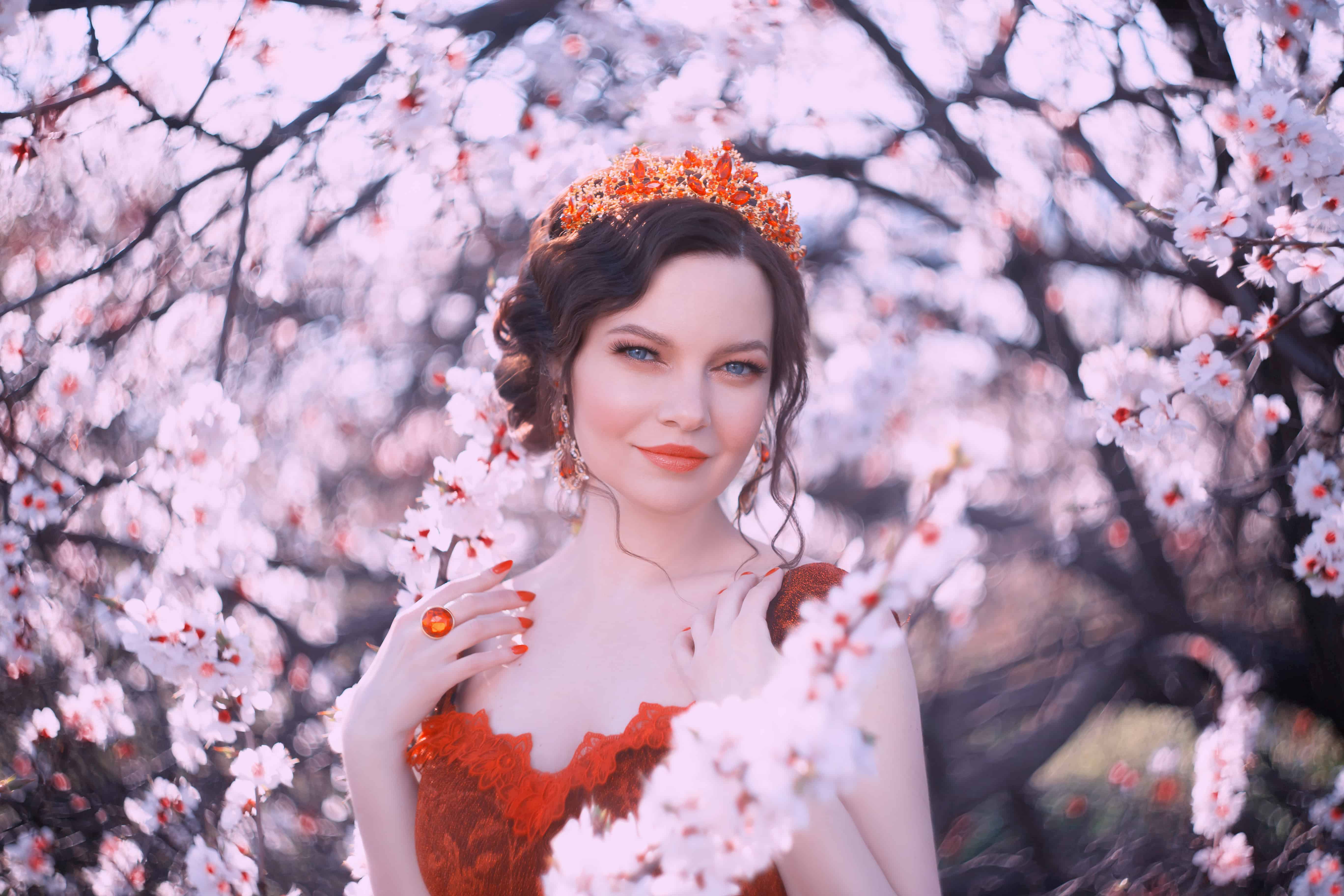 Queen of Spring walks in the blooming garden, a portrait photo of a pretty woman with dark hair and a golden crown on her head, the royal greatness, a red lady with bright blue eyes in sakura flowers