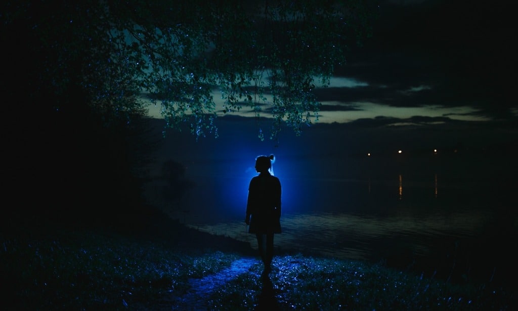 Silhouette of a young woman standing alone in the dark near the lake.