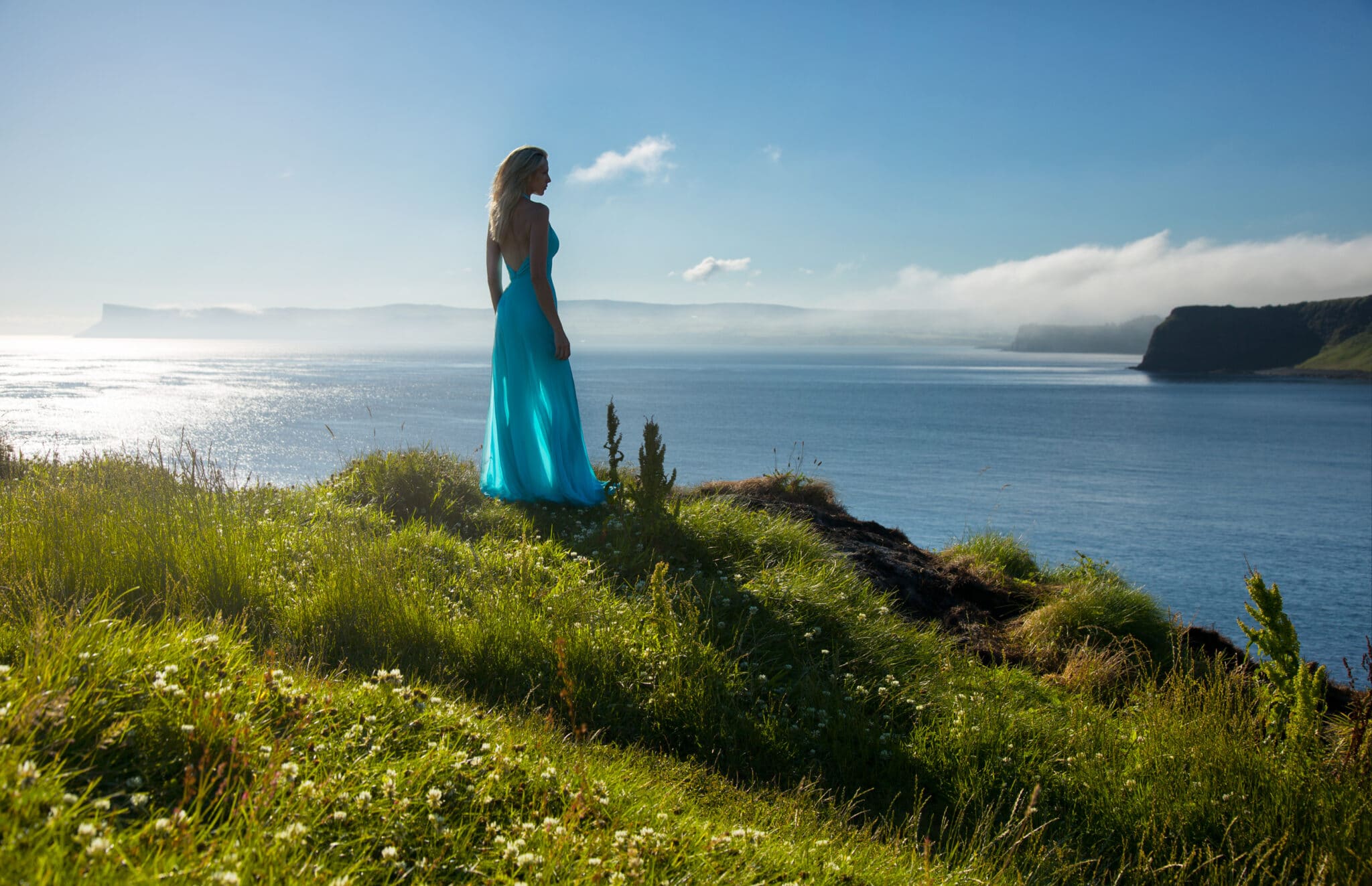 long haired blonde woman stands in long turquoise dress at the top of a coastal hill