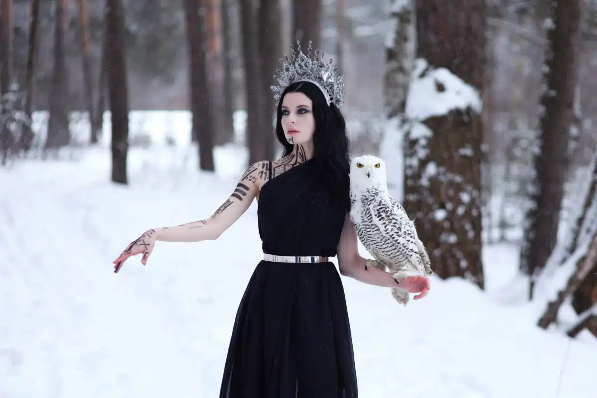 Beautiful brunette woman with body art in crown and black long dress with polar owl on hand standing in winter forest