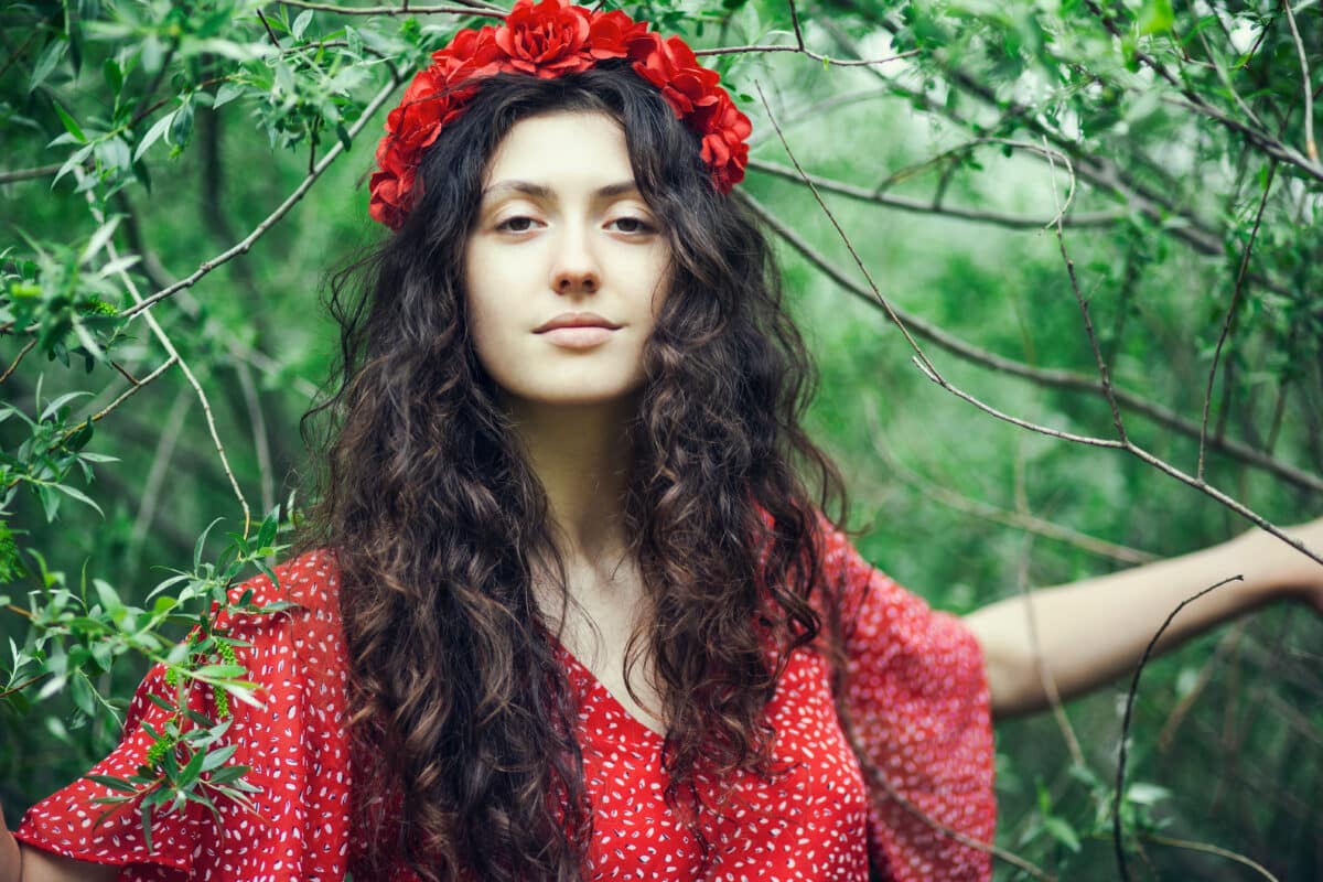 an enchanting lady in red with red flower crown standing in the woods