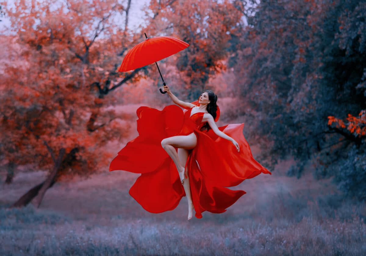 Beautiful elegant happy woman fly with umbrella. Red long sexy silk dress fabric fluttering in wind. goddess dancing joyful cheerful in air. Mystic autumn trees forest.