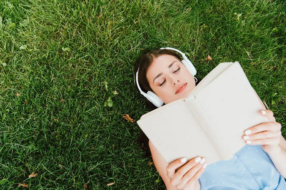 Attractive woman in wireless headphones lying on grass reading a book