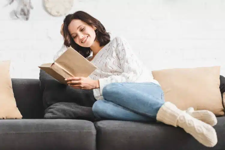 Pretty smiling young woman reading a book comfortably on a sofa at home.