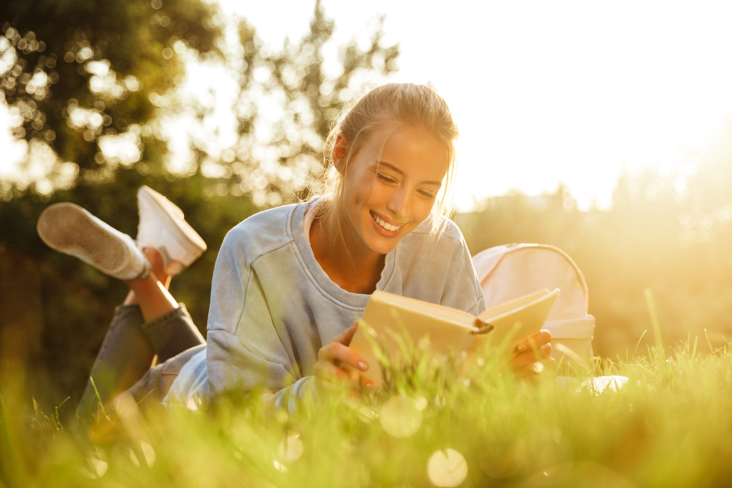 A smiling young girl reading a book on the grass outdoors.