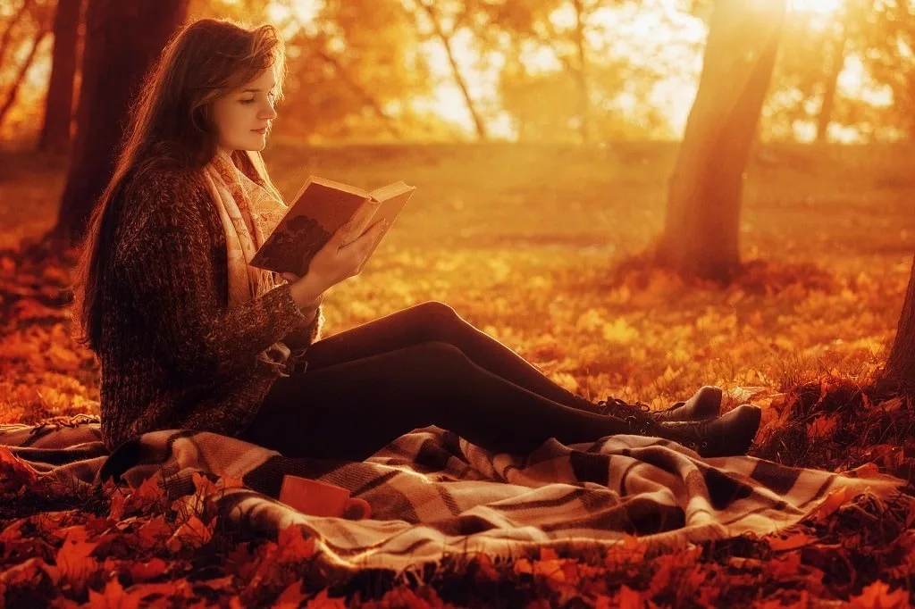Beautiful young brunette sitting on fallen autumn leaves in a park, reading a book