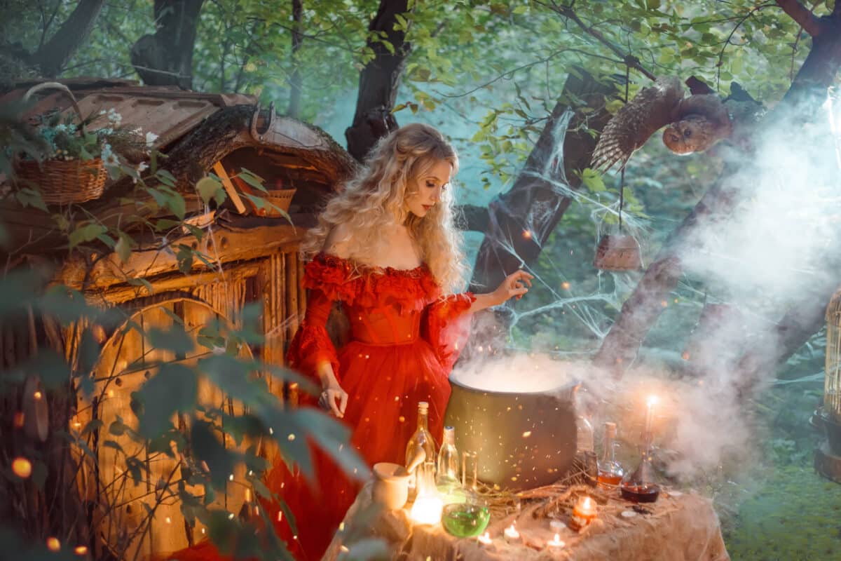 pretty young lady with blond curly hair above big magic cauldron with smoke and bottles with liquids, forest nymph in long bright red dress with loose sleeves prepares potion near wooden house
