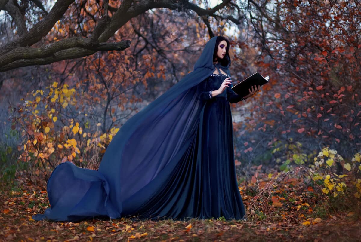 Art photography. Woman witch holds book in hands reads spell. Black clothes, cloak dress flies in wind. Fantasy girl elf in hood. Nature autumn forest, trees orange leaves. Holiday concept halloween