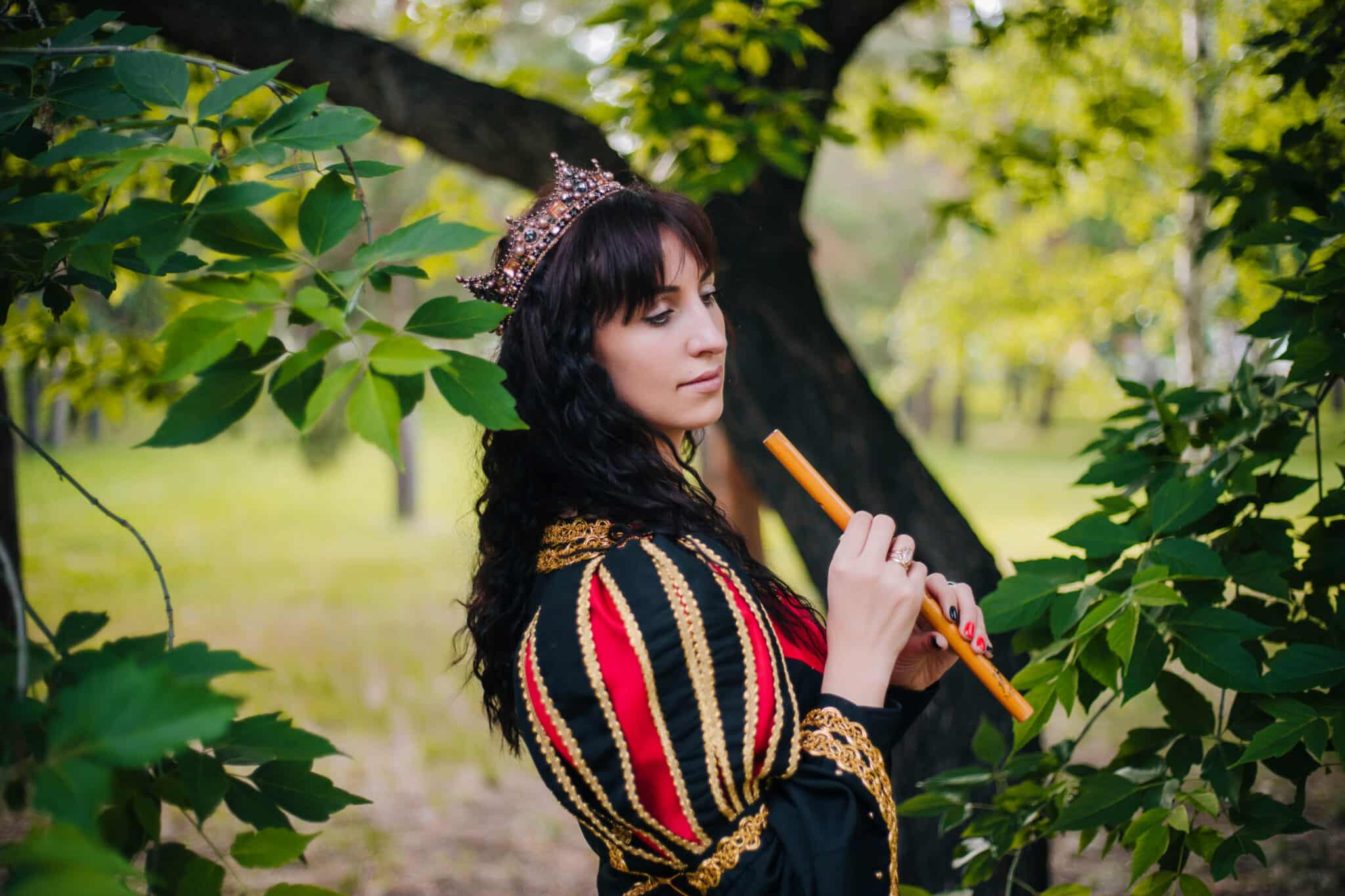 Queen with a flute in her hands. Beautiful girl in the crown plays on the pipe. Fantasy. Medieval black and red dress with gold embroidery.