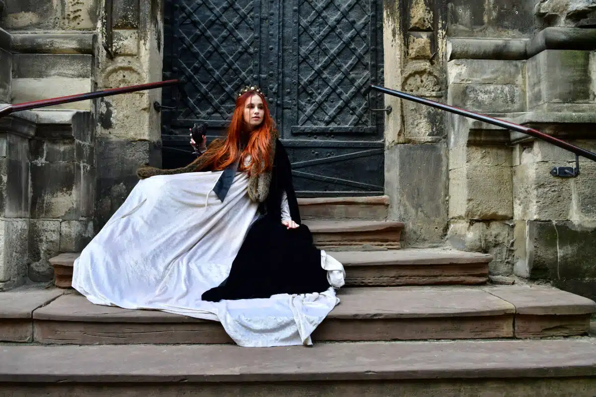 Red-haired princess in a white dress and a black veil with a crown sitting by the door on the concrete steps