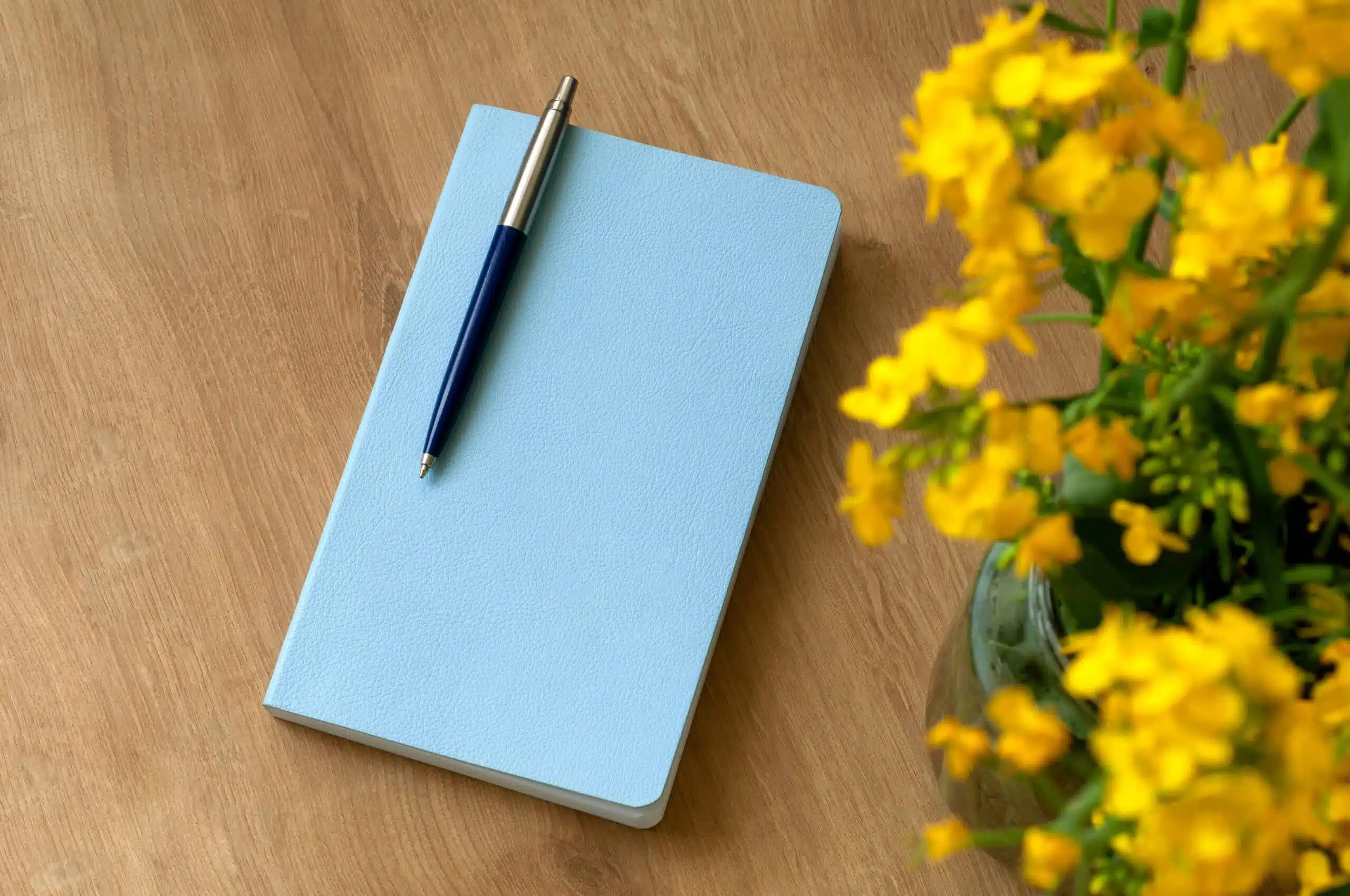 a blue notebook with a ballpoint pen lies on a table next to a vase with rapeseed flowers