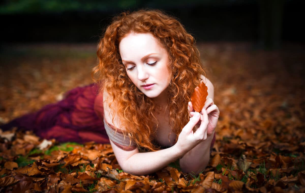 beautiful woman with long red wavy hair holding autumn leaf while lying on the ground with autumn leaves