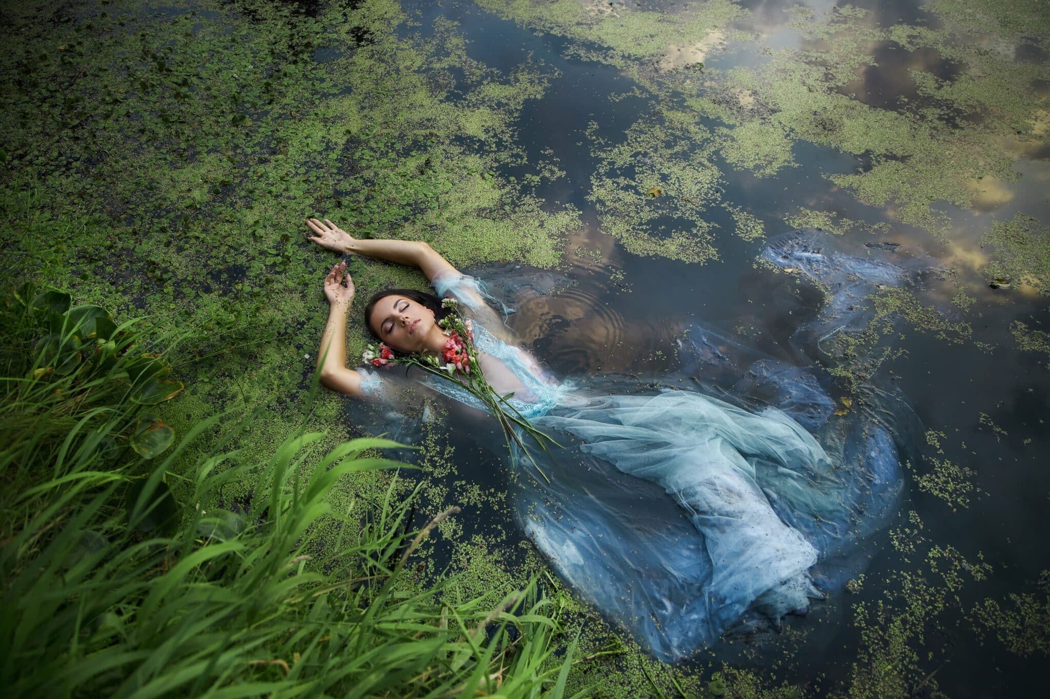 Art beautiful romantic woman lies in swamp in blue long dress with flowers.