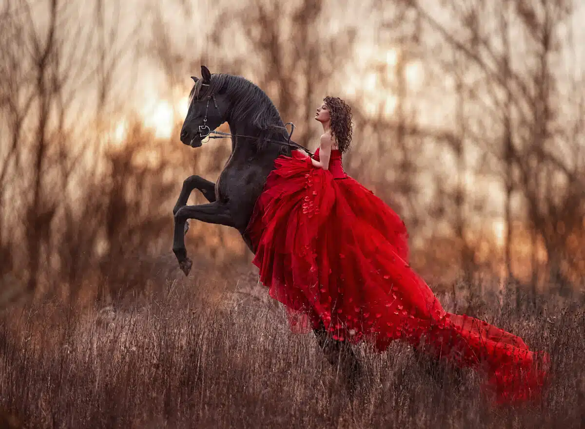 Beautiful curly-haired girl in a red dress riding a horse on its hind legs