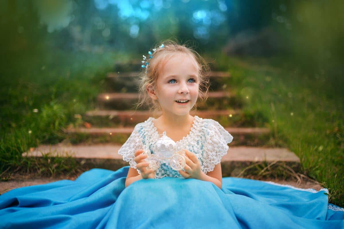 Little blond princess in a blue dress holds a small carriage in her hand