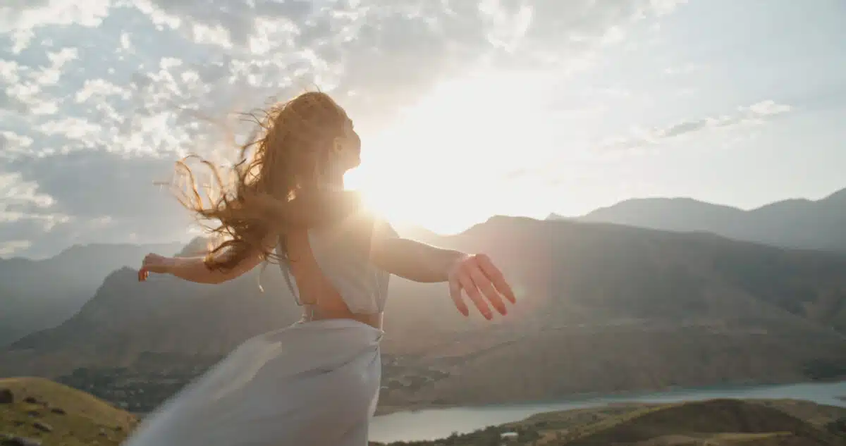 a woman in white dress standing on top of a mountain at sunset with raised hands while wind is blowing her dress and red hair