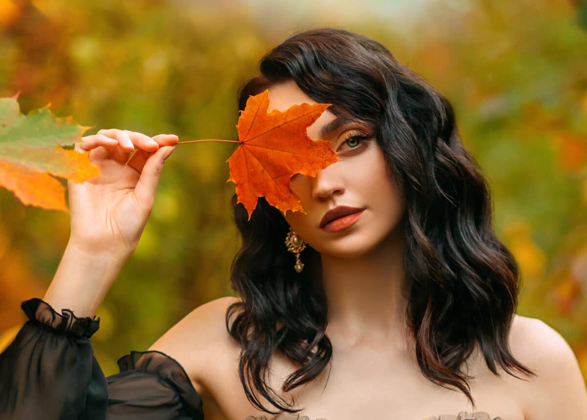 Fantasy girl princess holds red orange autumn fallen maple leaf in hands hides smiling happy face. Sexy woman queen looks at camera. black flowing hair. Yellow nature foliage trees. Art photo portrait