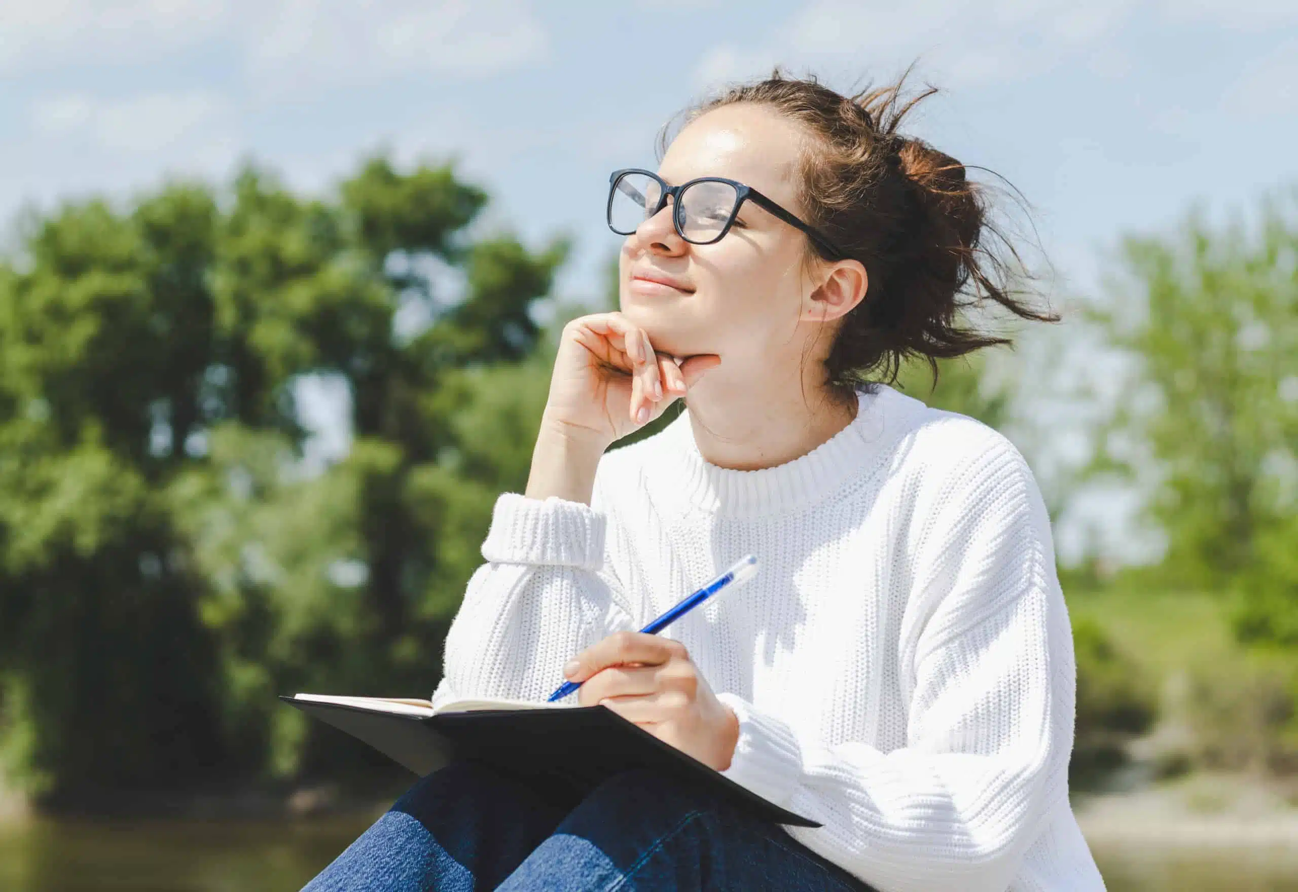 Young woman in glasses writes thoughts in notebook while relaxing outdoor.