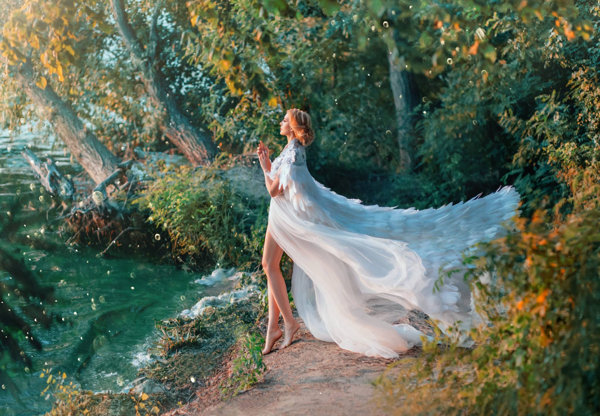 A woman is standing on the riverbank in a white dress and a raincoat with feathers. 