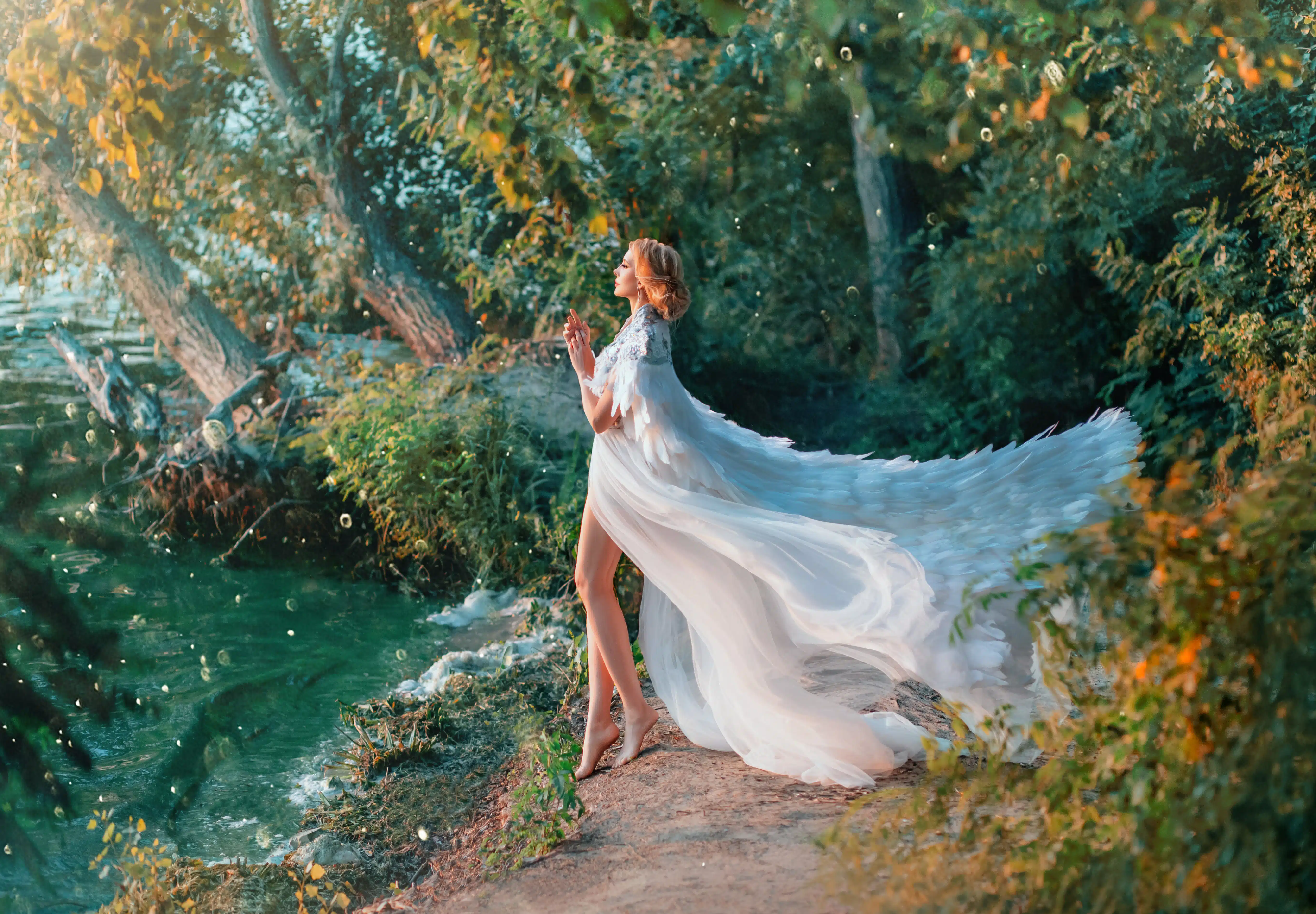 A woman is standing on the riverbank in a white dress and a raincoat with feathers. 