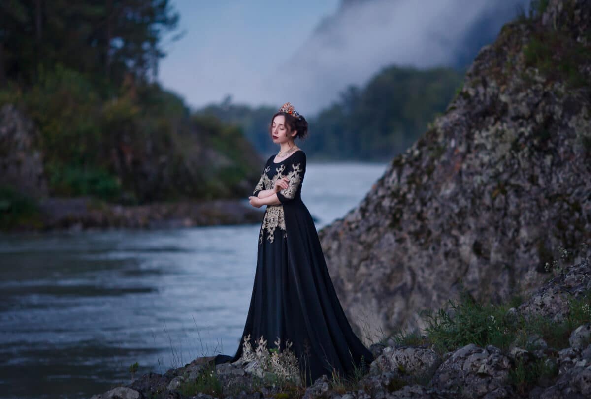 A girl in a black dress with a crown on her head stands on the banks of the river