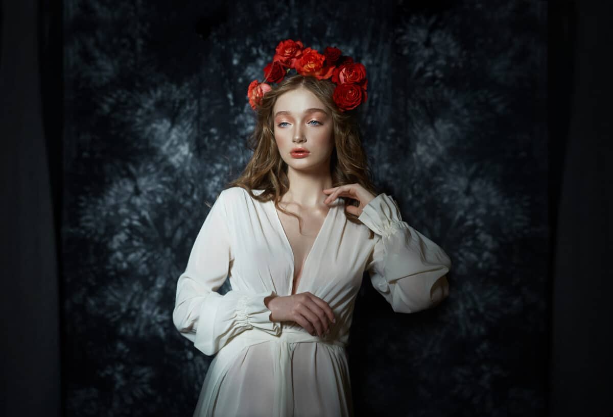 Romantic young woman in a white dress and a wreath of red rose flowers standing alone in the dark outdoors