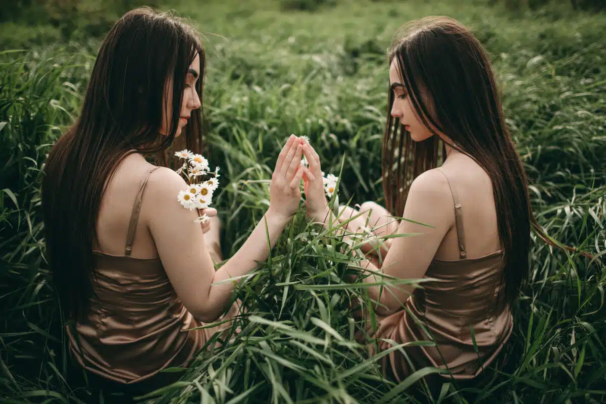 Beautiful young women twin sisters with long hair in a beige dress with chamomile flowers lie in the grass. Feelings, hugs.
