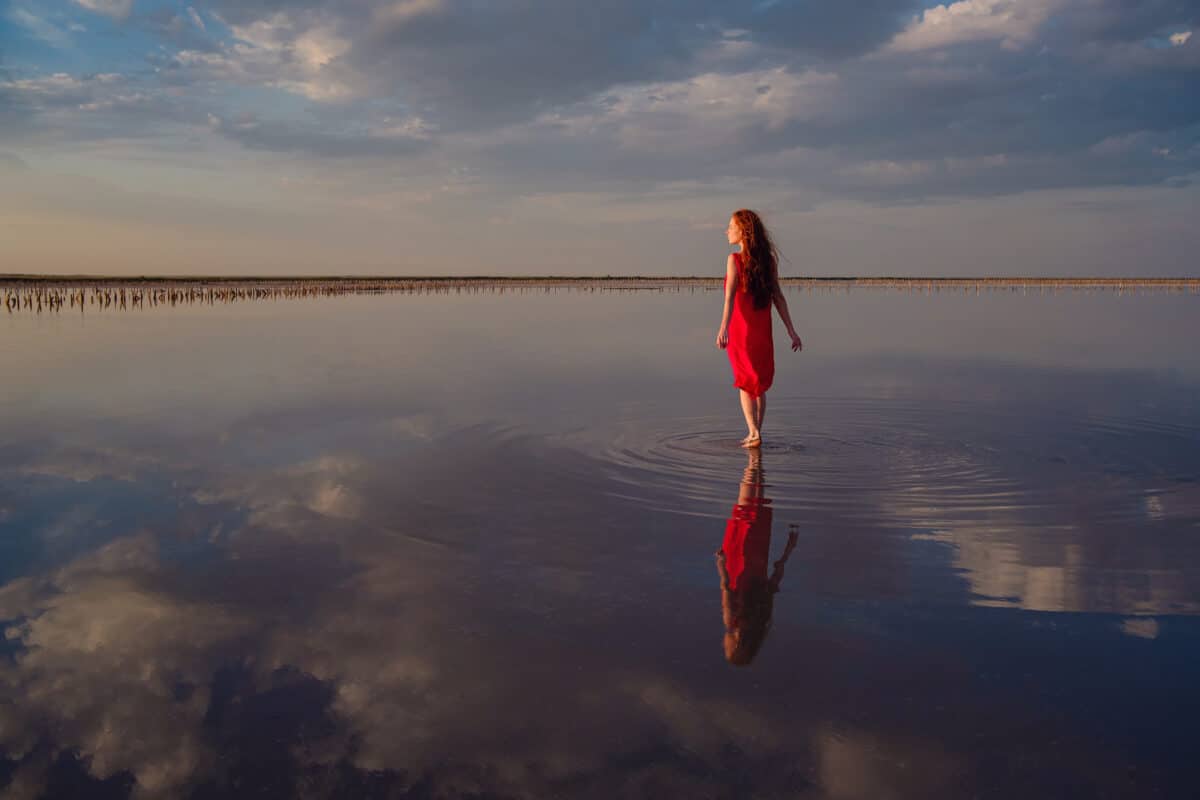 Elegant woman in red silky dress walking by a salt lake, her reflection in the water