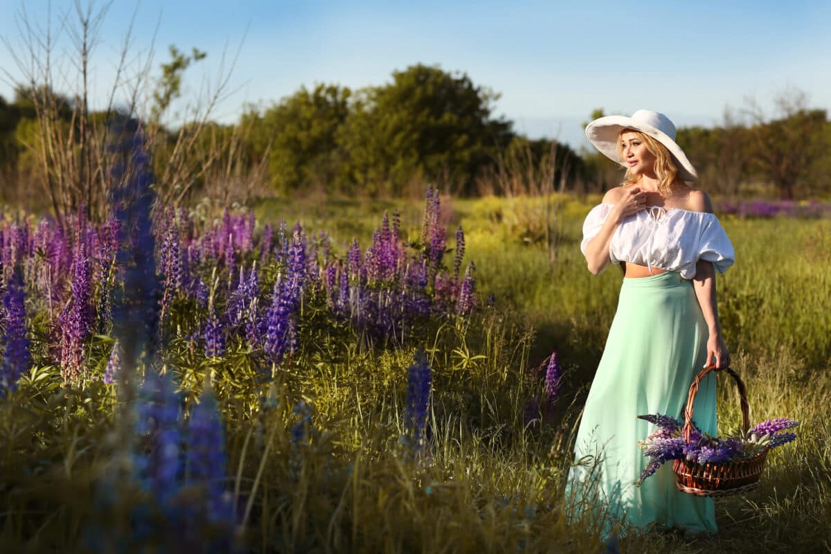 a beautiful blond lady in a white and green dress is picking violet flowers in the field