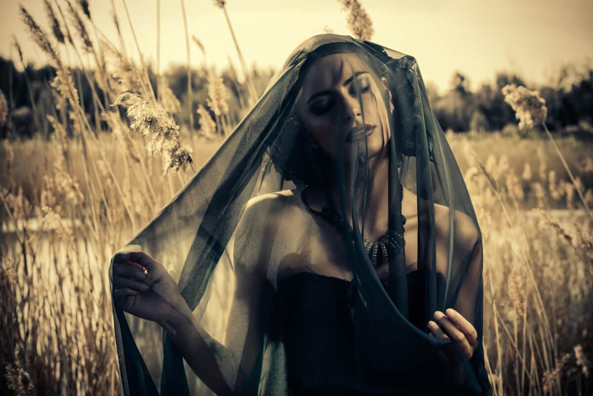 mysterious brunette woman in a long black dress and black veil standing among the reeds