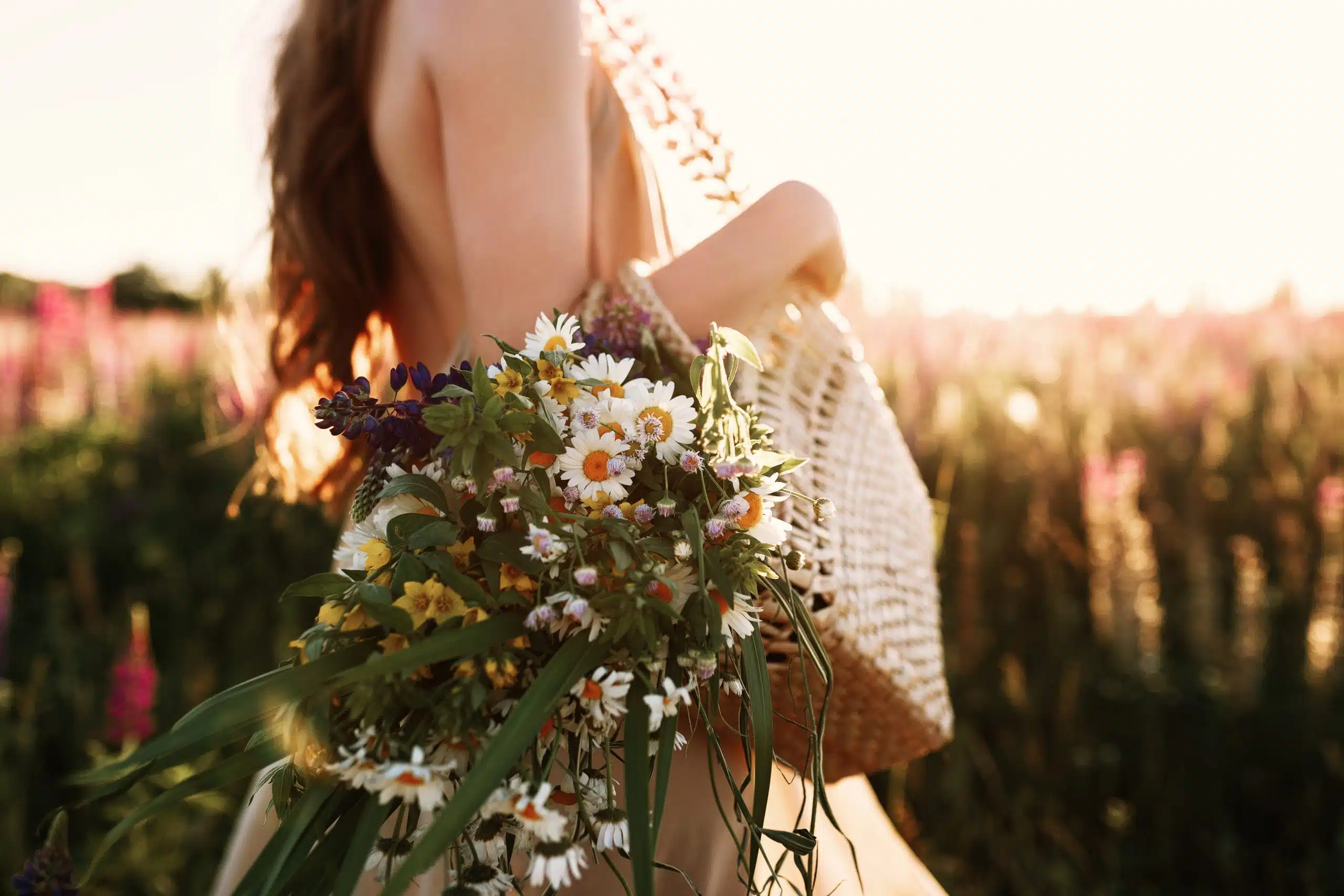 Woman holding wildflowers bouquet in straw bag, walking in flower field on sunset.