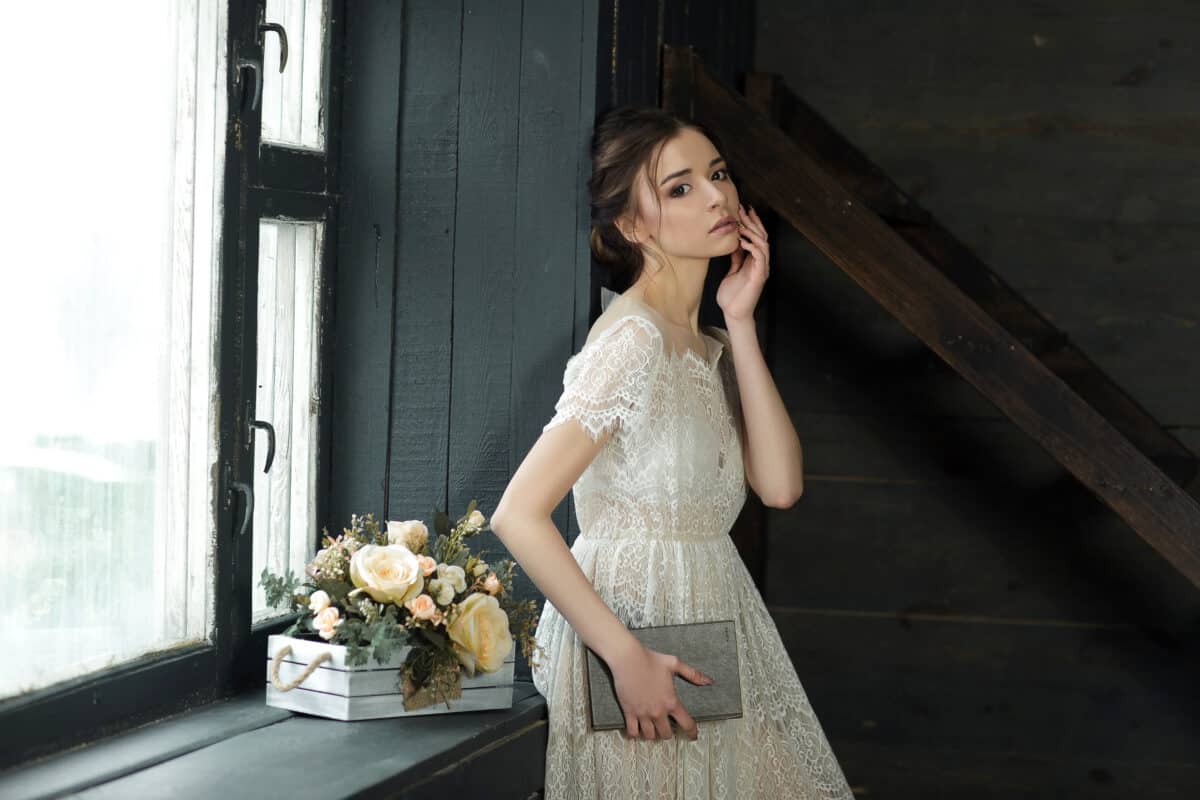 Pretty lady dressed in simple white dress with a book standing near the window
