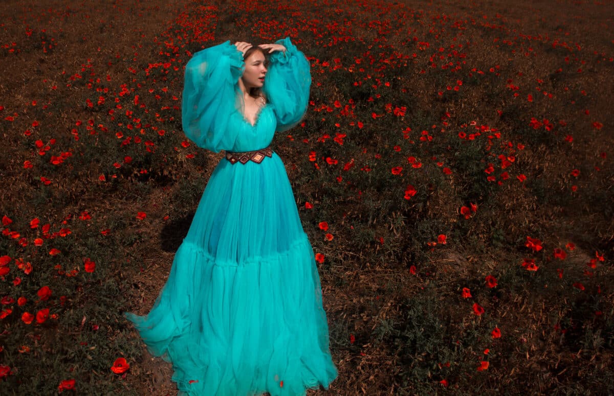 young blonde lady with long hair in huge blue princess dress in a field of poppies